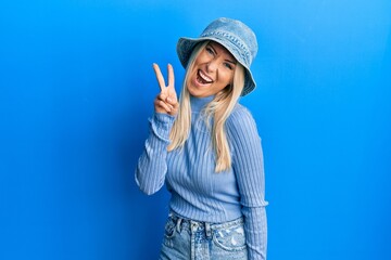 Young blonde woman wearing casual denim hat smiling with happy face winking at the camera doing victory sign. number two.