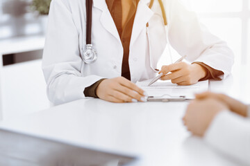 Unknown woman-doctor and female patient sitting and talking at medical examination in sunny clinic, close-up. Medicine concept