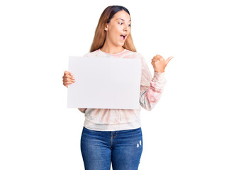 Beautiful young woman holding blank empty banner pointing thumb up to the side smiling happy with open mouth