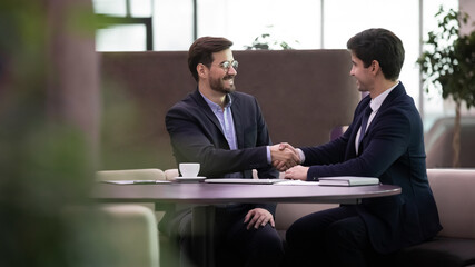 Smiling young Caucasian businessmen shake hands get acquainted greet at meeting in office. Happy male colleagues or business partners handshake make agreement or close deal. Partnership concept.