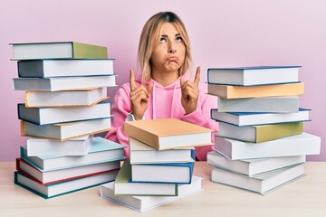 Young caucasian woman sitting on the table with books pointing up looking sad and upset, indicating direction with fingers, unhappy and depressed.