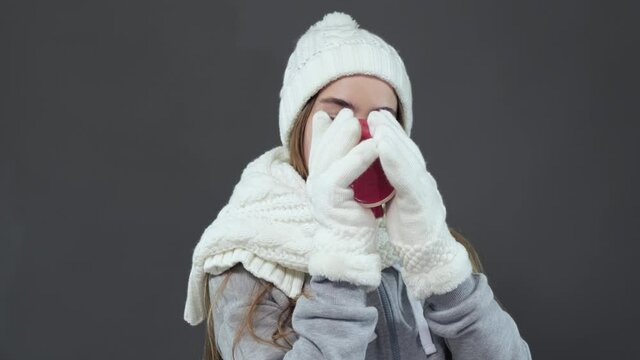 Portrait of a young beautiful girl in a white winter hat and gloves enjoying a delicious coffee while standing in the studio.