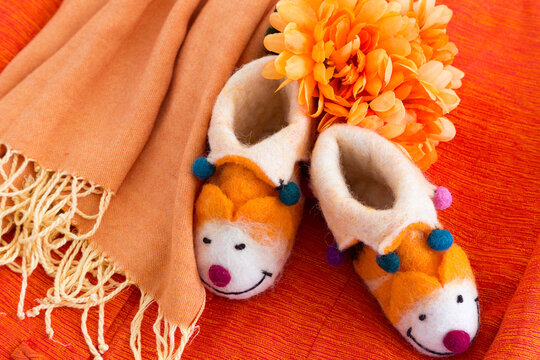 Orange Themed Flat Lay Still Life With Background Of Raw Silk Jacket, Cute Felt Baby Slippers, Silk Flowers And Fringed Scarf