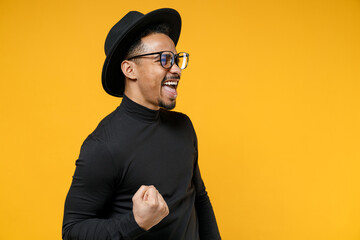 Young smiling happy overjoyed fun african american man 20s wearing stylish black hat shirt eyeglasses do winner gesture clench fist celebrating say yes isolated on yellow background studio portrait.