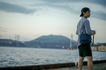 Portrait Asian man standing on the port and looking out to the sea with the twilight sky of sunset and bokeh lights of the city in background