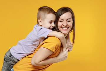 Happy young woman have fun with cute child baby boy 5-6-7 years old in violet t-shirt stand behind hug kiss Mommy little kid son together isolated on yellow background studio Mother's Day love family