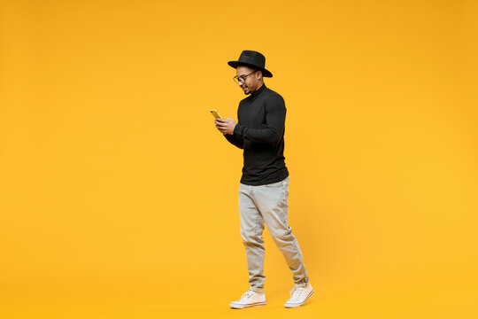 Full Length Side View Young Friendly Smiling Happy African Man In Stylish Black Shirt Hat Eyeglasses Using Mobile Cell Phone Chat In Social Network Walk Isolated On Yellow Background Studio Portrait