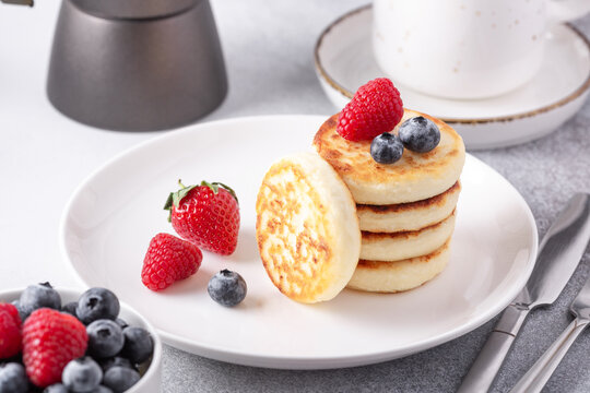 Cottage cheese pancakes with fresh berries, cup of coffee and coffee maker on the table. Tasty breakfast food - Image