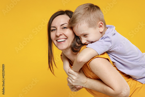 Happy young woman have fun with cute child baby boy 5-6-7 years old in violet t-shirt stand behind hug kiss Mommy little kid son together isolated on yellow background studio Mother's Day love family