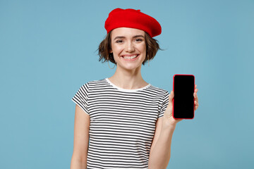 Young happy european woman 20s with short hairdo wearing french beret red hat striped tshirt hold mobile cell phone with blank screen workspace area isolated on pastel blue background studio portrait