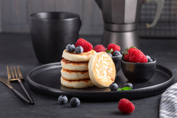Cottage cheese pancakes with fresh with fresh strawberry and blueberries on dark ceramic plate. Cup of coffee and coffee maker - Image