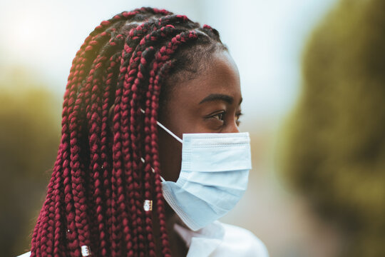 Portrait with a shallow depth of field and a selective focus on a black beautiful lady with braids of red color, looking aside while standing outdoors with a surgical virus protective mask on her face