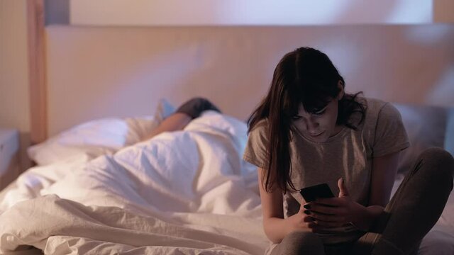 Internet anxiety. Night insomnia. Social media. Disturbed woman using mobile phone sitting on bed with sleeping husband late in home bedroom.