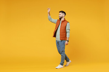 Full length young smiling happy confident smiling cheerful fun caucasian man 20s wearing orange vest mint sweatshirt walking going waving hand greeting isolated on yellow background studio portrait - obrazy, fototapety, plakaty