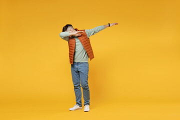 Full length young caucasian fun man in orange vest mint sweatshirt glasses doing dab hip hop dance hands move gesture youth sign hiding cover face isolated on yellow color background studio portrait