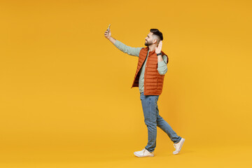 Fototapeta na wymiar Full length young smiling friendly man 20s in orange vest mint sweatshirt doing selfie shot on mobile phone waving hand greet say hello isolated on yellow background studio. People lifestyle concept.