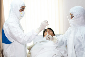 Two medical technicians wearing full protective equipment collected samples from Asian young man lying in a hospital bed, the operation of the coronavirus mobile testing unit.