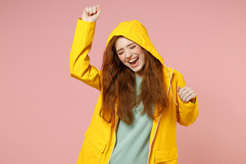 Redhead young smiling fun woman 20s in yellow waterproof raincoat outerwear dancing enjoy isolated on pastel pink color background studio portrait. Outdoors lifestyle wet fall weather season concept