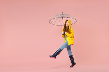 Full length side view redhead young woman in yellow waterproof raincoat hood outerwear hold transparent umbrella walking go isolated on pink background studio Outdoor lifestyle wet weather concept