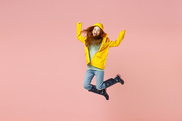 Full length redhead excited young woman in yellow waterproof raincoat outerwear jump high do winner gesture isolated on pastel pink background studio Outdoor lifestyle wet fall weather season concept.