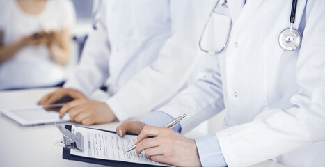 Unknown woman-doctors at work with patient at the background. Female physicians filling up medical documents or prescription while standing in hospital reception desk, close-up. Health care concept