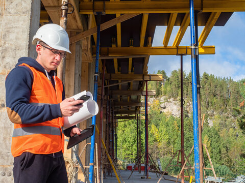 Engineer Calling Someone. Architect-engineer On Background Of A House Under Construction. Engineer At A Construction Site. Man In Construction Worker's Uniform. Concept - Erection Of A Country Hotel