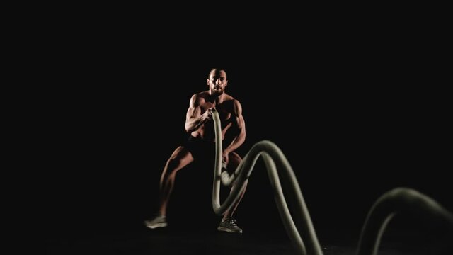 Athletic man training with battle ropes on black background. Slow motion