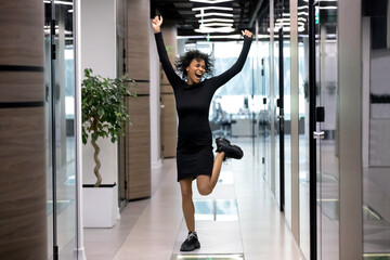 Excited African American woman worker have Friday fun or party enjoy job promotion or achievement. Overjoyed millennial biracial female employee dance celebrate work results or success in office.