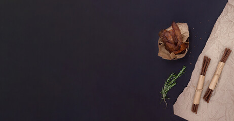 Horizontal banner with dried meat sticks, wooden bowl with jerky and rosemary on craft paper on black background with copy space. Spiced meat for beer.