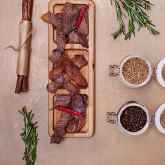 Jerky snacks on wooden board, meat sticks, peppercorns in glass jars and rosemary on light brown background. Top view. Dried spiced meat for beer.