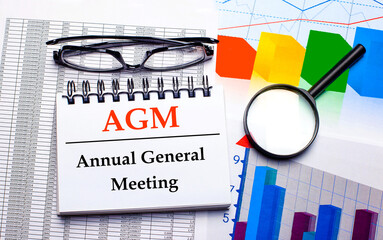 On the desktop are glasses, a magnifying glass, color charts and a white notebook with the text AGM Annual General Meeting. Business concept. View from above