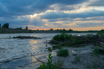 The sun's rays fall through the clouds on the river bank in the evening, in front of the green bushes.