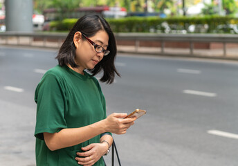 Asian middle-aged woman looking at smartphone in hand, wearing eyeglasses, standing on street sidewalk. Concept for waiting and using smartphone to track her uber taxi in city.