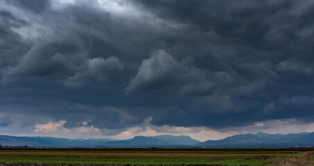 Dramatic storm clouds over empty green agricultural fields, blue mountains at the horizon line.