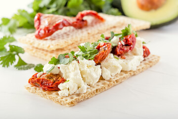 Crispy low-calorie wheat crackers with curd cheese and sun-dried tomatoes on a background of avocado and greens on a white wooden table. Close-up