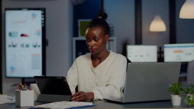 African manager woman using laptop and tablet in same time working overtime in business start-up office. Busy multitasking Busy employee analysing financial statistics overworking writing, searching.