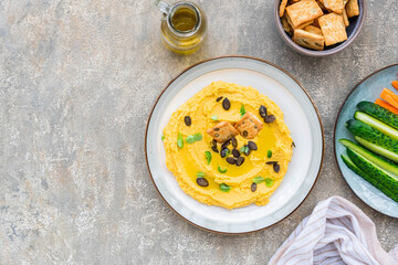 Healthy snack, chickpea hummus with pumpkin or carrots and pumpkin seeds on a ceramic plate on a gray concrete background. Served with savory crackers.