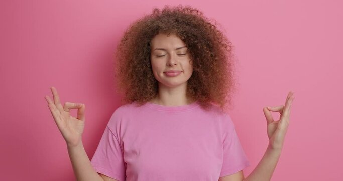 Curly haired Caucasian woman meditates with closed eyes holds mind under control practices yoga makes zen gesture dressed in casual t shirt isolated over pink background. Relaxation calmness