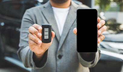 Close up businessman in suit his hands showing or giving car key for customer getting new car and showing screen of smartphone. Buy sell rent car concept.