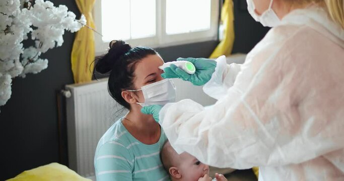 Doctor in a protective suit measures the temperature of a  girl holding a baby in her arms.