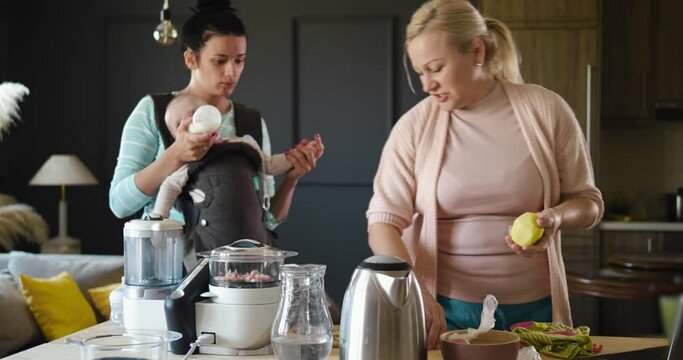 Multi-generational family in the kitchen, preparing baby food. Young mother holds the baby in a kangaroo and gives him milk to drink, the grandmother helps her daughter to prepare lunch