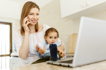Beautiful business mom is talking on the mobile phone and taking notes while spending time with her cute baby boy at kitchen home office. Working mother concept,Laptop on table
