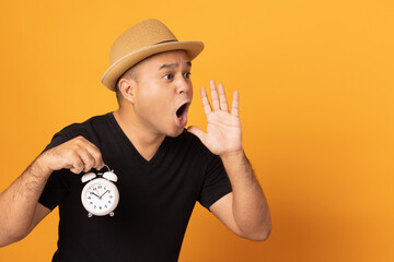 The young man covered his mouth with his hand and shouted loudly. Blowing loud announcements on isolated yellow background with copy space. He holding Big Clock.