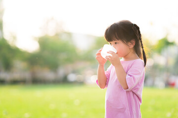 Adorable Asian child girl are drinking fresh water from plastic glasses. Green natural background. In the summer or spring. Side view of kid is 4 year old wearing pink shirt thirsty.
