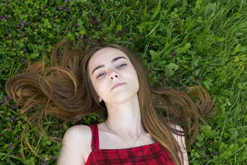 A beautiful thin young girl teenager in a red plaid sundress dress lies on the green grass, on the ground in summer.