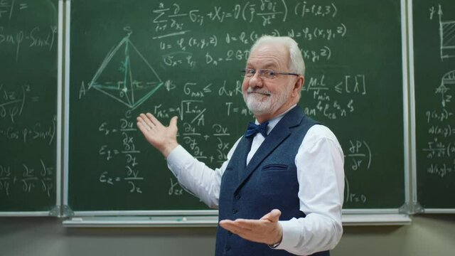 Intelligent senior teacher in university classroom standing looking at camera and smiles, backdrop blackboard with mathematical formulas. Happy gray haired professor. Education and studying concept