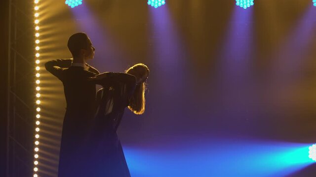 Elegant couple of ballroom dancers dancing waltz elements bouncing and holding hands. A man and a woman waltz in a dark studio amid bright blue spotlights and smoke. Close up.