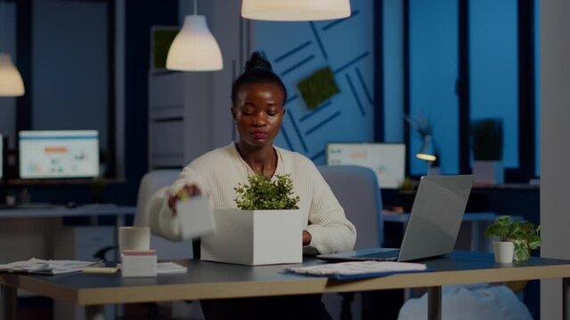 Sad african fired businesswoman packing things in box being dismissed from job late at night. Overworked unemployed woman closing laptop and leaving workplace office in midnight