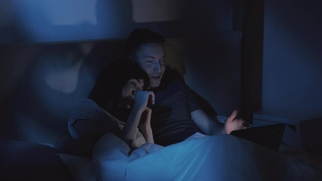 Night online shopping. Happy couple. Family order. Excited wife choosing gift with husband using laptop in bed late in dark home bedroom with blue light.