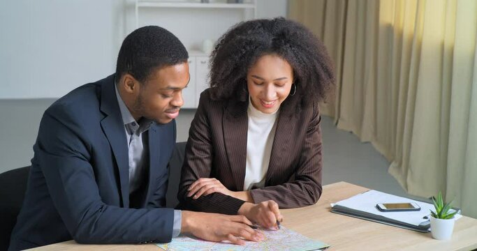 Two business colleagues Afro American man and woman in formal suits sitting at table looking at map searching place for construction building comany architect points shows schematic plan, top view